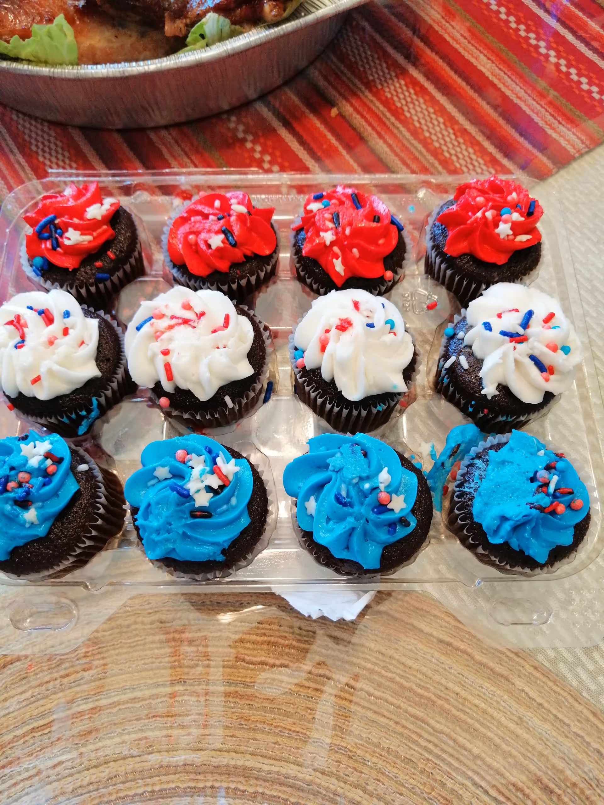 A clear plastic tray of chocolate cupcakes topped with red, white, and blue frosting and sprinkles on a table.
