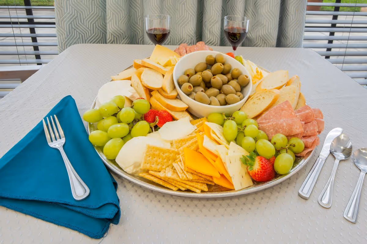 A large platter on a table with an assortment of green grapes, strawberries, crackers, various cheeses, sliced bread, olives in a bowl, and slices of deli meat. Two glasses of red wine are placed behind the platter. A blue napkin with a fork is on the left side of the table, and three spoons are on the right side. The table is covered with a white tablecloth and there are patterned curtains and window blinds in the background.