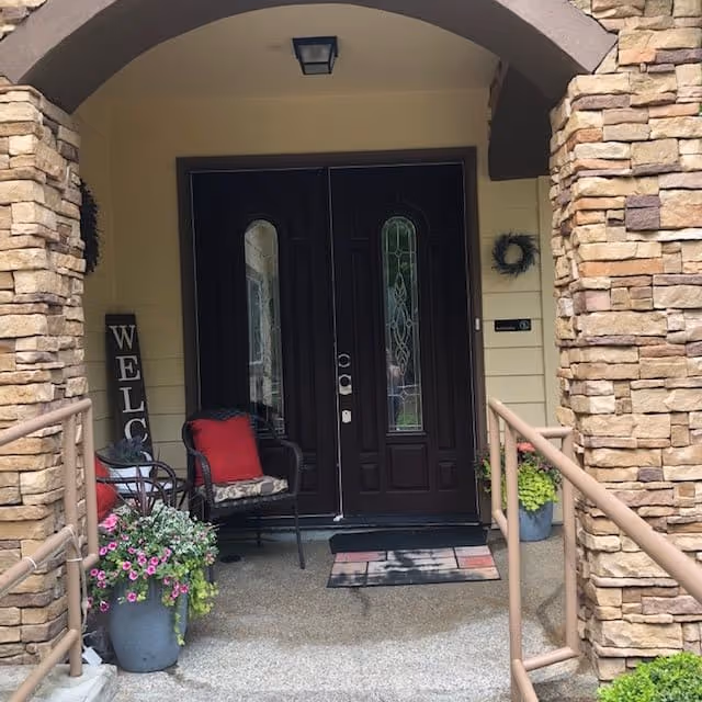 Entrance to a building with double dark wooden doors featuring decorative glass panels. The entrance is framed by stone pillars and an archway. There is a black chair with a red cushion and a small table with a red cushion on the left side. Two potted plants with flowers are placed near the entrance. A vertical wooden sign with the word 'WELCOME' is leaning against the wall on the left side. A doormat is placed in front of the doors.