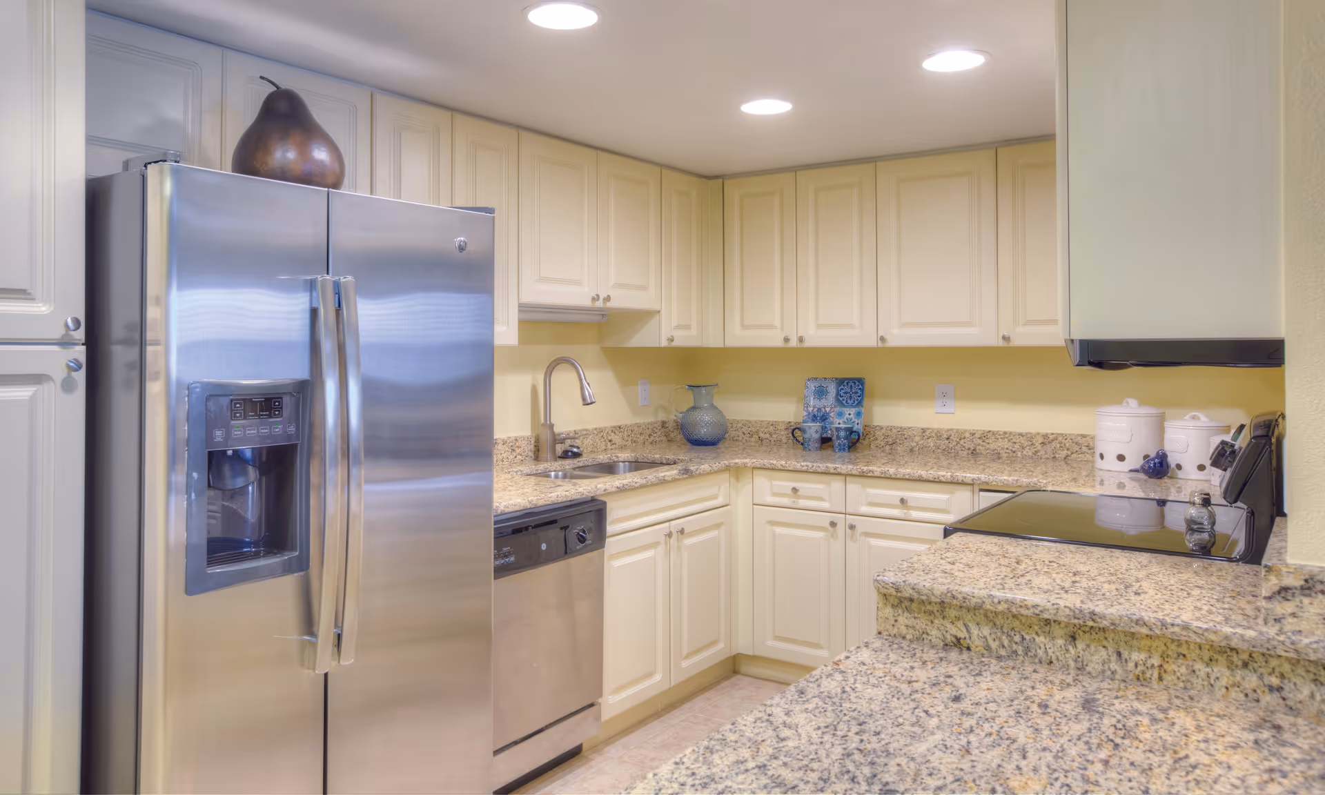 Bright kitchen with cream-colored cabinets, granite countertops, stainless steel refrigerator with water dispenser, dishwasher, and electric stove. Decorative items including a large pear sculpture on top of the refrigerator, a blue glass pitcher, two blue patterned mugs, and white ceramic containers are placed on the countertops.