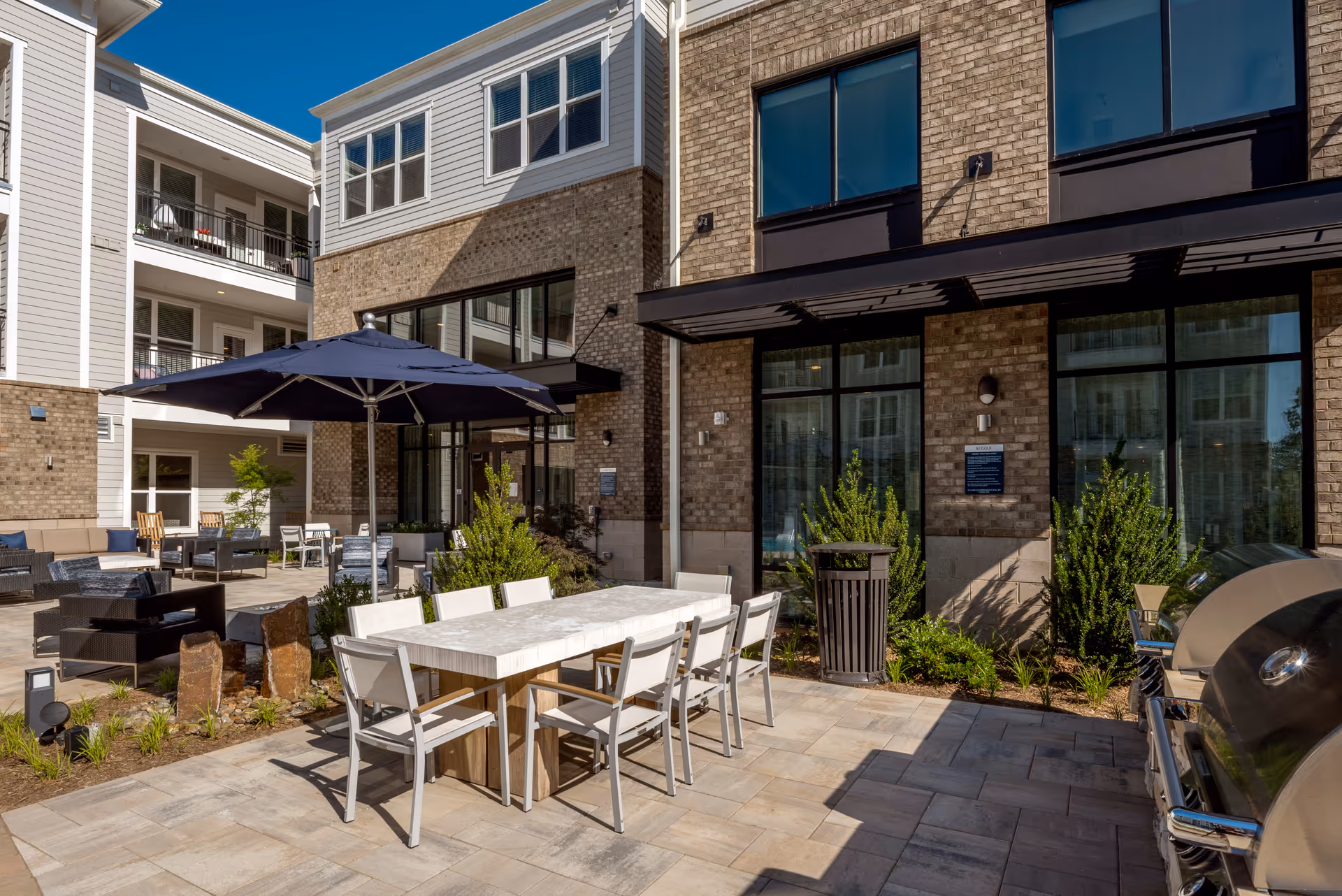 Outdoor patio area at a senior living facility with a rectangular dining table surrounded by white chairs, a large blue umbrella providing shade, lounge seating, plants, and a barbecue grill. The building exterior features brick and siding with large windows.