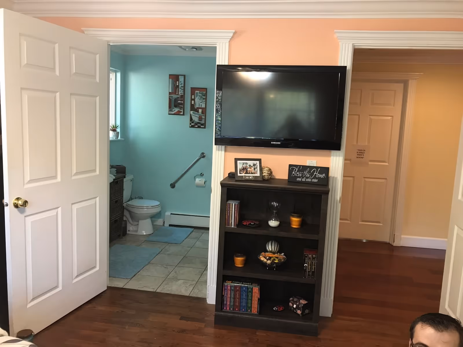 View of an interior space showing a wall-mounted flat-screen TV above a dark wooden bookshelf filled with books, decorative items, and framed photos. To the left, an open door reveals a bathroom with light blue walls, a toilet, and a window. To the right, a hallway with closed doors is visible. The walls in the main room are painted peach, and the floor is wooden.