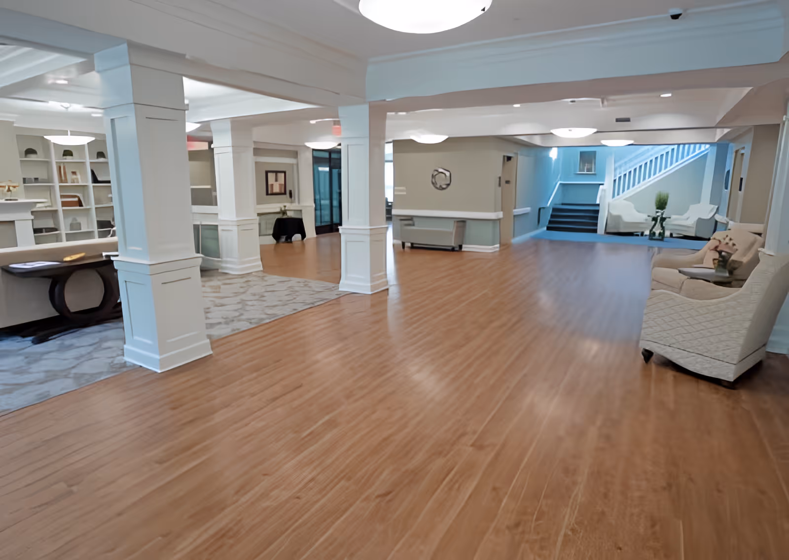 Spacious interior common area with wooden flooring, white pillars, and seating arrangements including sofas and chairs. There are built-in shelves with decorative items on the left side and a staircase leading to an upper level in the background.
