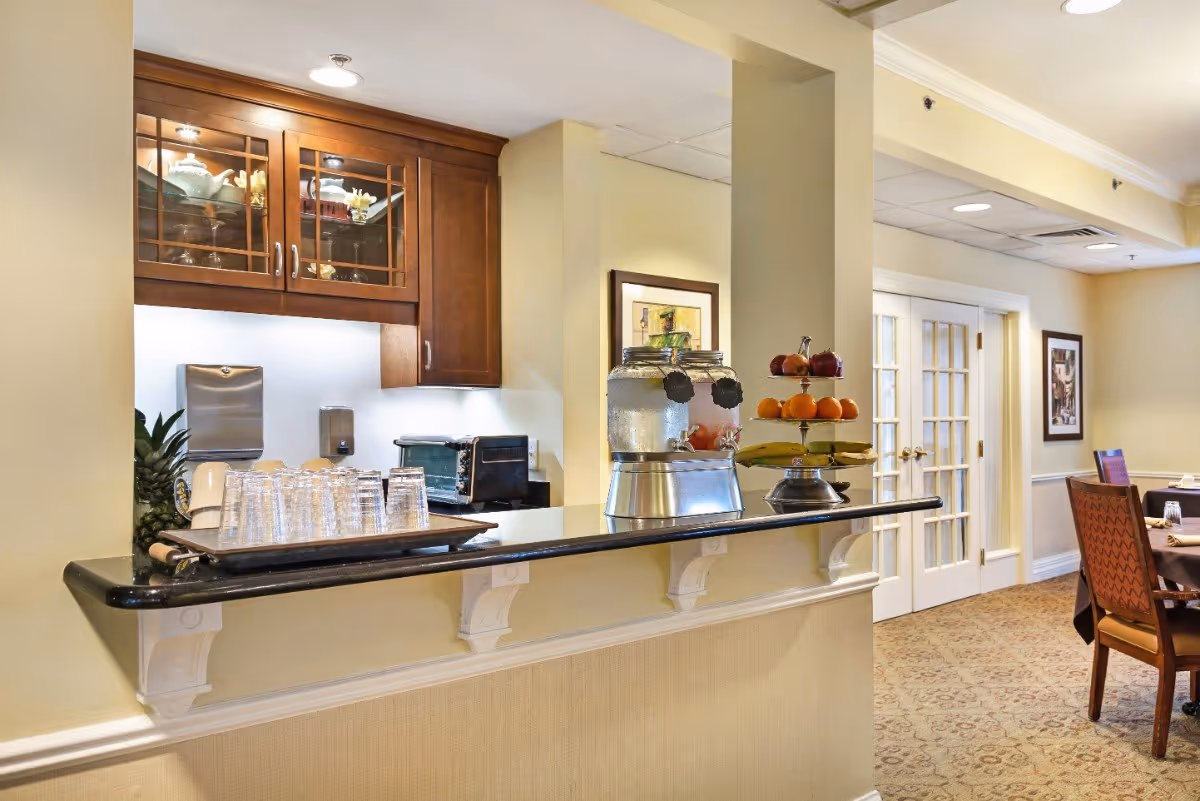 Interior view of a senior living facility dining area with a counter displaying a tray of glasses, two beverage dispensers, and a tiered fruit stand. Behind the counter are wooden cabinets and a toaster oven. The dining area has carpeted floors, a table with chairs, framed artwork on the walls, and French doors in the background.