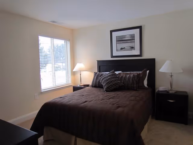 A bedroom with a neatly made bed featuring a dark brown comforter and several patterned pillows. There are two matching dark nightstands on either side of the bed, each with a lamp. A framed black and white picture hangs above the headboard. A window with white blinds allows natural light into the room.