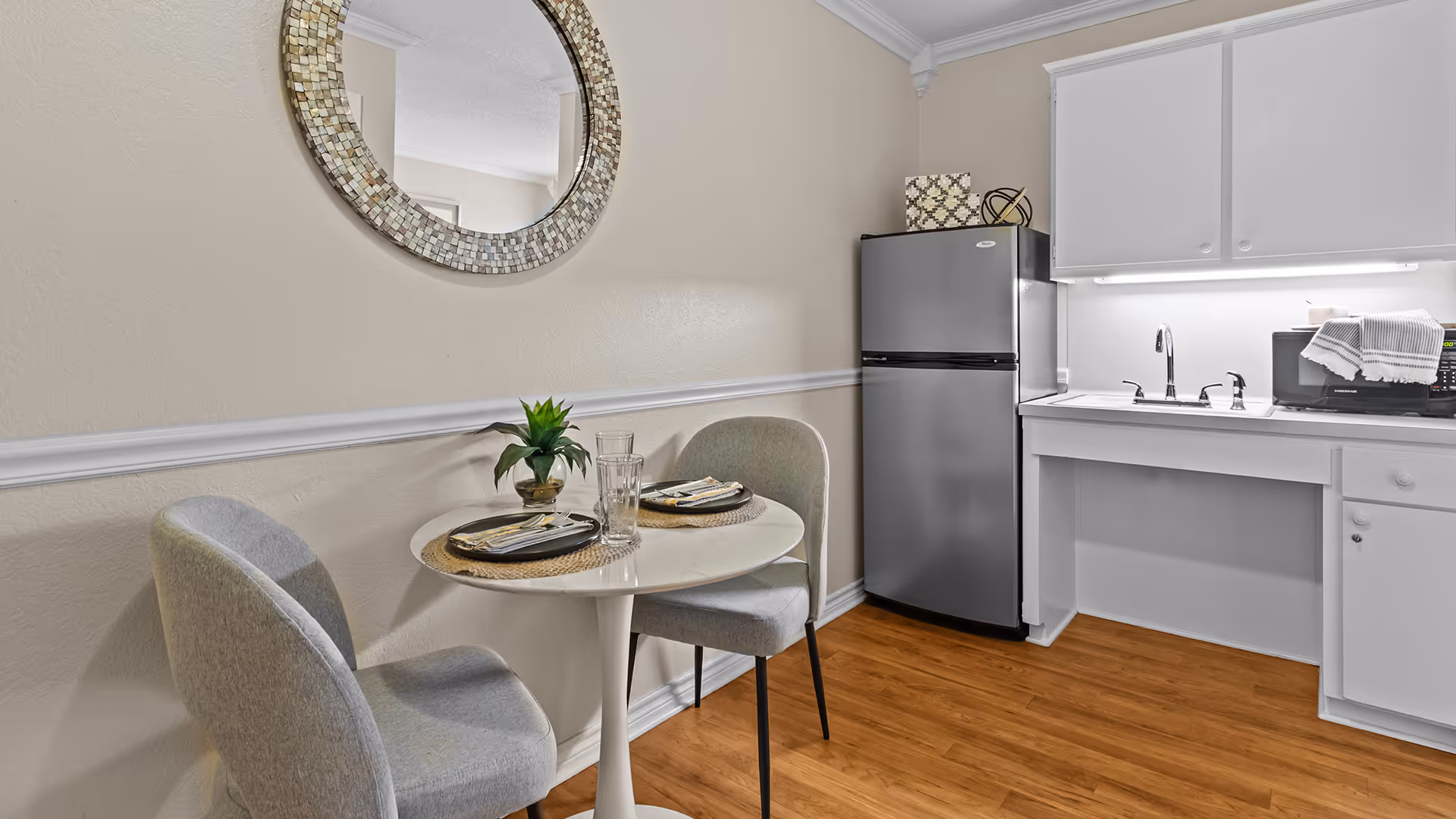 Small kitchen area with a round white table set for two with plates, napkins, and glasses. Two gray upholstered chairs are positioned at the table. A decorative round mirror hangs on the beige wall above the table. To the right, there is a stainless steel refrigerator, white cabinets, a sink, and a microwave on the countertop. The floor is wood.