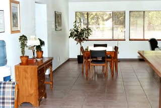 A bright interior room with tiled flooring featuring a wooden table with four chairs near large windows. There is a wooden sideboard with potted plants and a lamp on the left side, and a water cooler next to it. The room has white walls and framed pictures hanging on them.