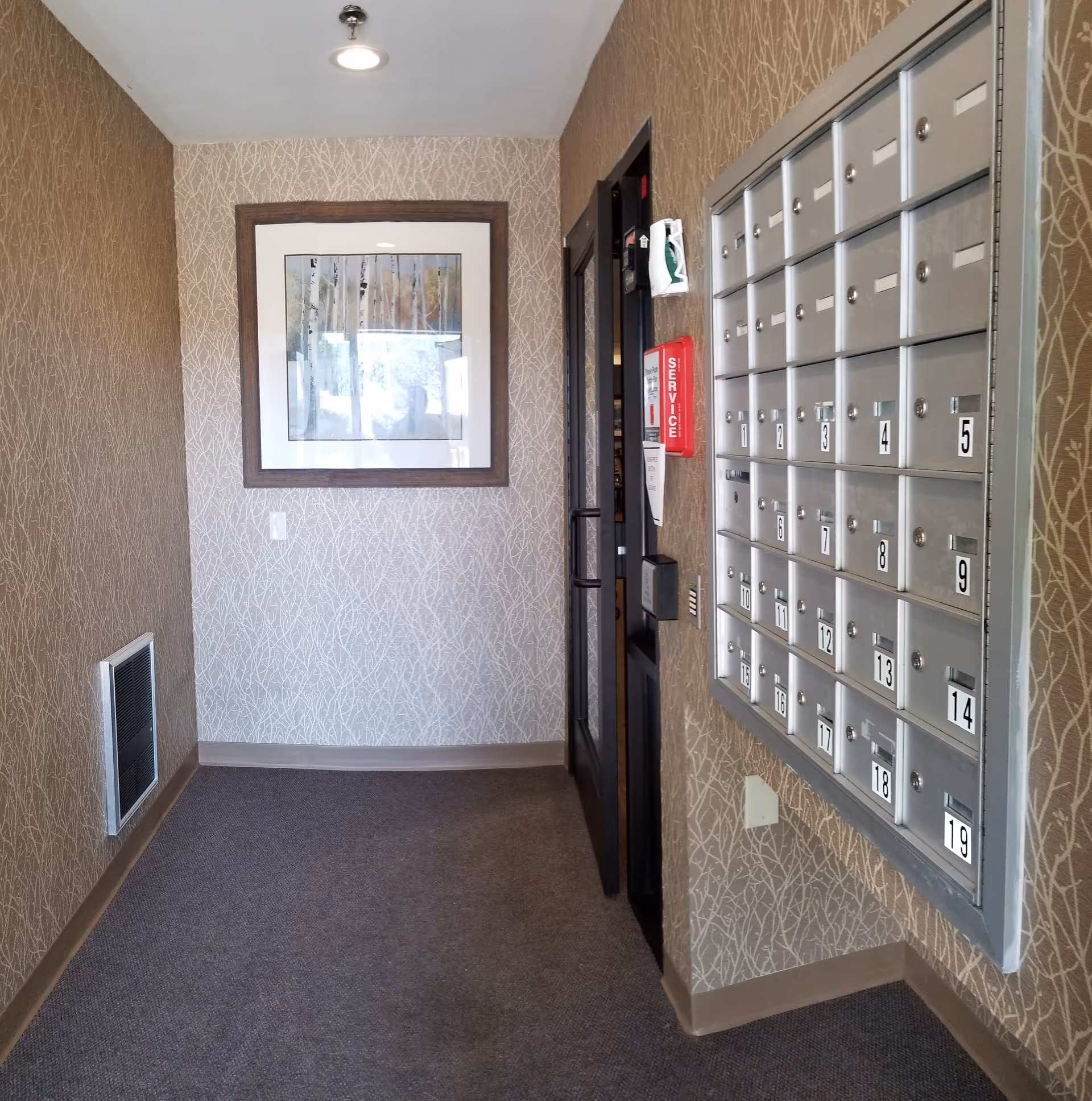 Interior hallway area with a wall-mounted set of numbered mailboxes on the right, a framed picture of trees on the far wall, a fire alarm service box, and a door with glass panels.