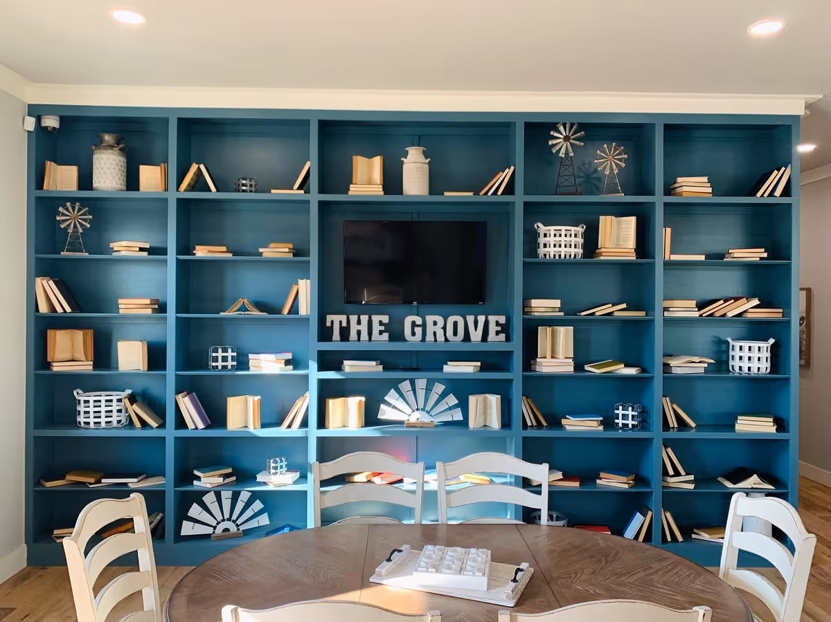 Teal built-in bookcase filled with books and decor, a TV and 'THE GROVE' sign above a round dining table with chairs.