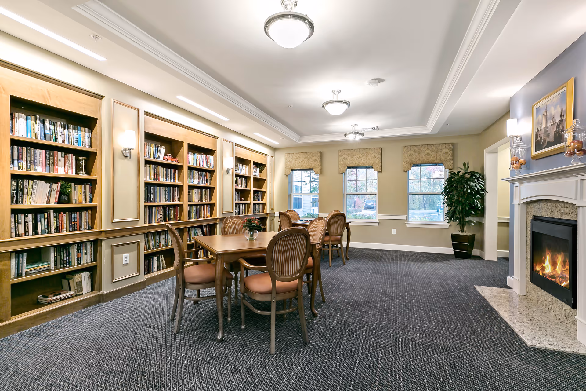 A cozy library room with built-in wooden bookshelves filled with books along one wall. There are two wooden tables with cushioned chairs arranged around them. The room has three windows with decorative valances, a fireplace with a lit fire, a potted plant in the corner, and framed artwork above the fireplace. The carpet is dark patterned, and the ceiling has recessed lighting and ceiling fixtures.