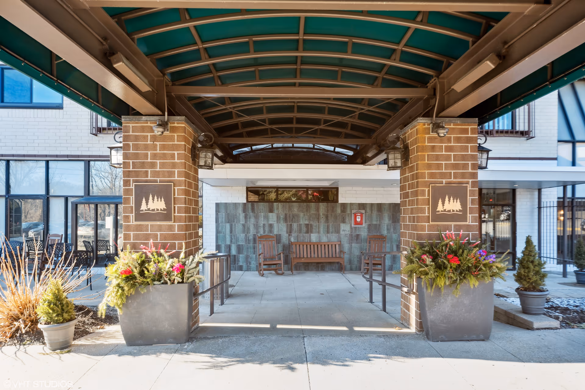 Covered entrance to a senior living building with brick columns, potted planters and benches under a green awning.