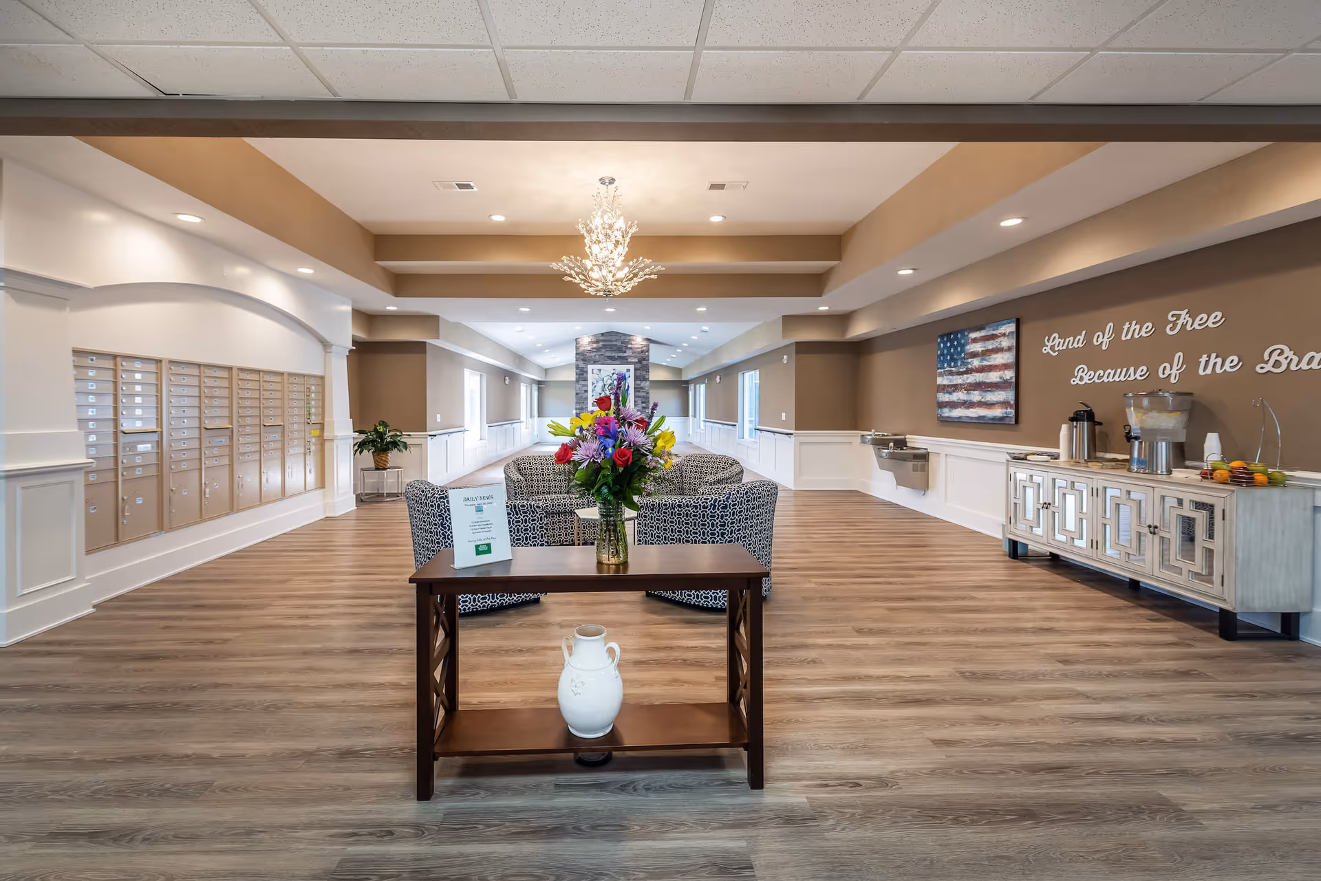 A spacious and well-lit interior common area with wood flooring and beige walls. In the center, there is a small wooden table with a vase of colorful flowers and a sign. Behind the table, there are patterned armchairs arranged around a stone fireplace. On the left wall, there are multiple mailboxes. On the right wall, a sideboard holds a coffee station and a water dispenser, with a patriotic painting of the American flag and the words 'Land of the Free Because of the Brave' displayed above.