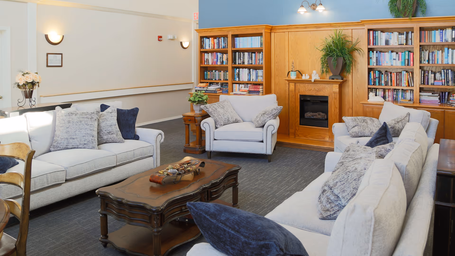 A cozy living room area with light gray sofas and armchairs adorned with various pillows. A wooden coffee table sits in the center with decorative items on it. Behind the seating area is a wooden built-in bookshelf filled with books and a small fireplace in the middle. The room has soft lighting and a plant on top of the bookshelf.