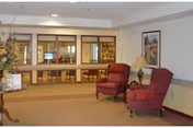 A quiet interior common area with two red upholstered armchairs, a side table with a lamp, a framed picture on the wall, and a large windowed partition showing another room with tables and chairs.