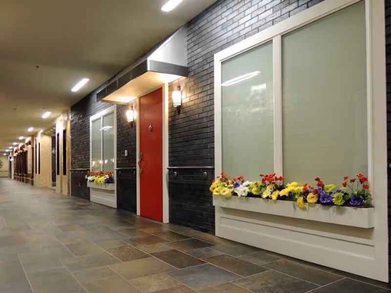 Indoor corridor with tiled floor, black brick walls, a red door and window boxes filled with colorful flowers.