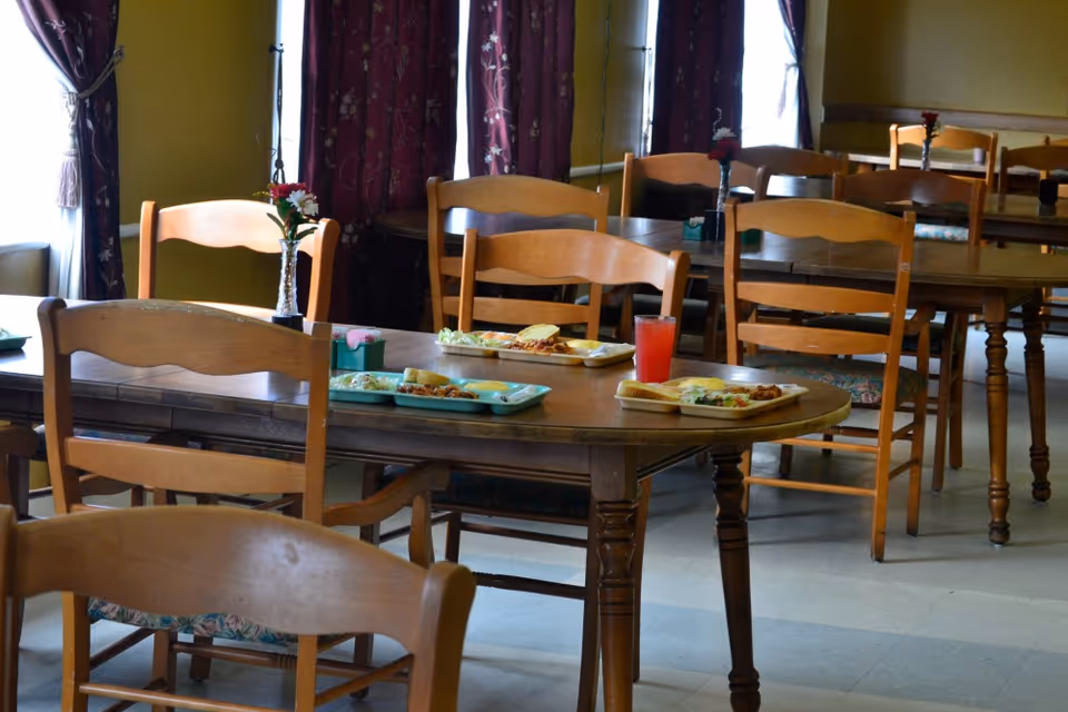 Communal dining room with wooden tables and chairs, meal trays and a red drink on one table.