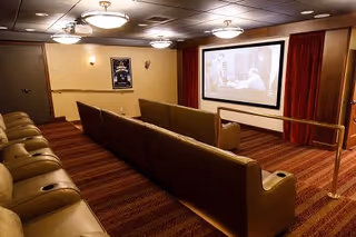 A small theater room with two rows of brown leather seats facing a large screen showing a black and white movie. The room has warm lighting with ceiling lights and wall sconces, red curtains on either side of the screen, and a patterned carpet.