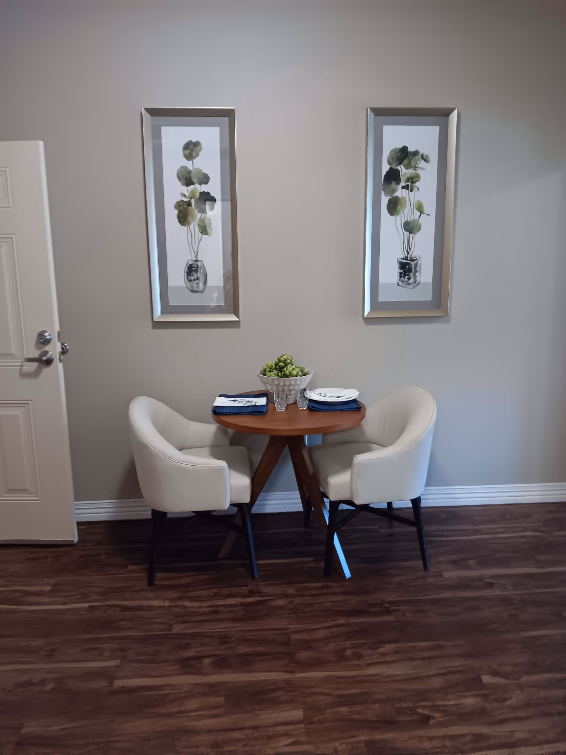 A small round wooden dining table set for two with white cushioned chairs on a dark wood floor. The table has two place settings with plates, napkins, and glasses, and a decorative bowl with green artichokes in the center. Two framed botanical prints hang on the light gray wall behind the table, and a white door is partially visible on the left side.