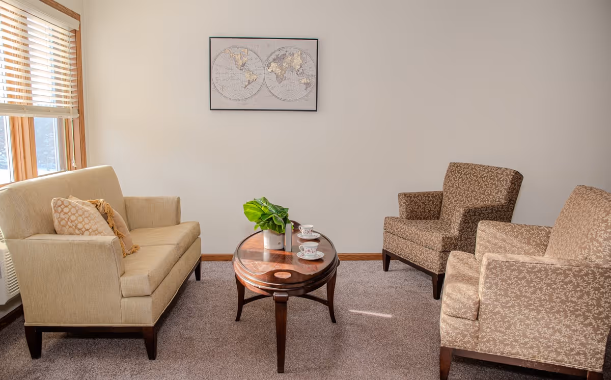 A cozy living room area with a beige loveseat on the left, two patterned armchairs on the right, and a round wooden coffee table in the center with a small potted plant and two teacups. A framed world map hangs on the plain light-colored wall, and sunlight streams in through a window with wooden blinds on the left.
