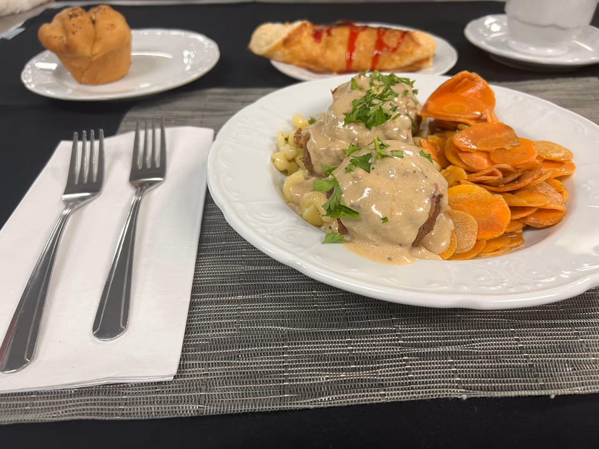 A plated meal consisting of two pieces of meat covered in creamy sauce and garnished with parsley, served with sliced cooked carrots and pasta. On the table, there are two forks on a white napkin, a bread roll on a small white plate, a croissant with ketchup on a small white plate, and a white cup and saucer in the background.