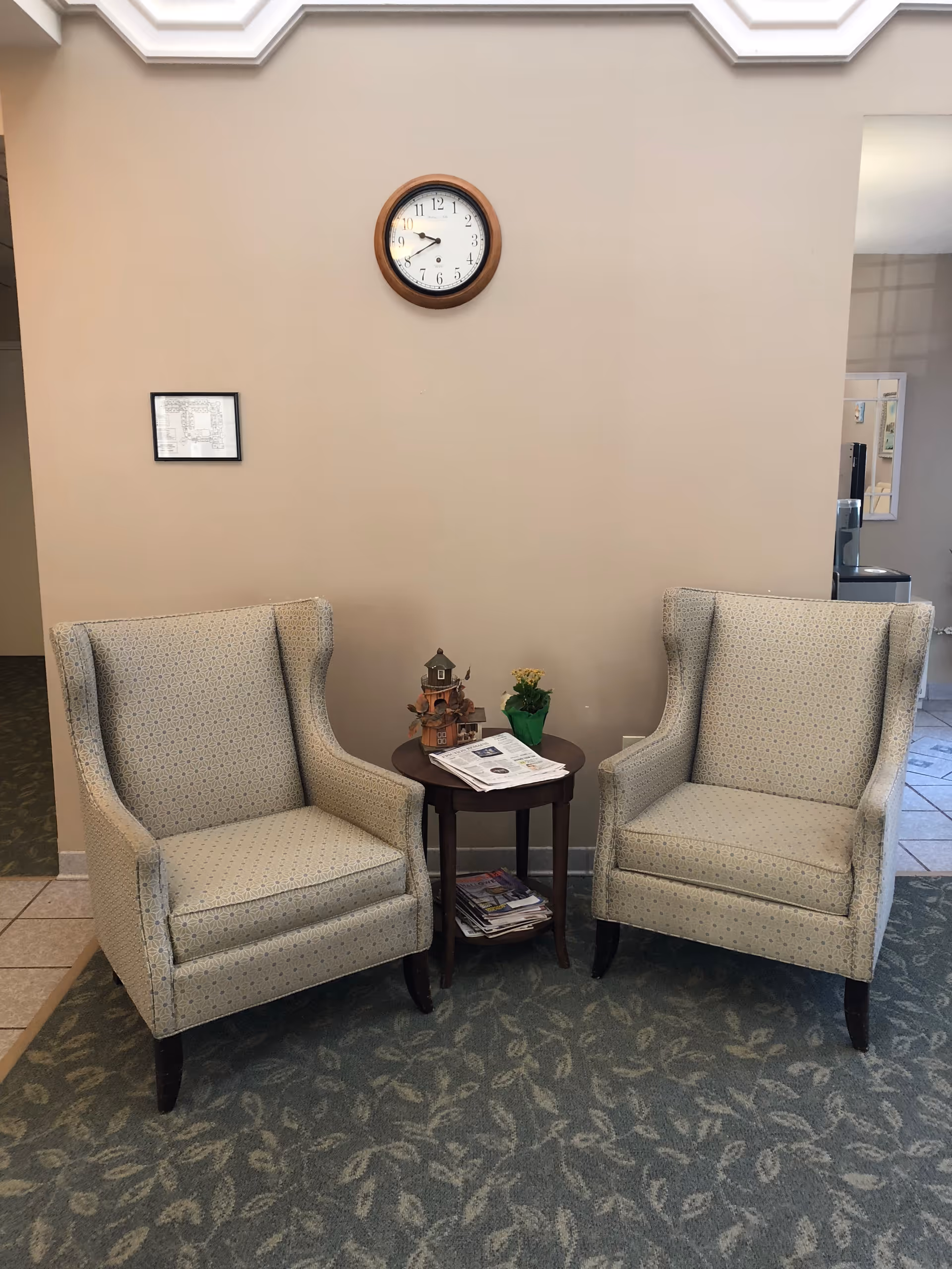 Two beige patterned armchairs with wooden legs are positioned on a green carpet with a leaf pattern. Between the chairs is a small round wooden table holding a miniature house decoration, a small potted plant, and some newspapers. Above the chairs on the beige wall is a round clock showing the time as 9:45, and a small framed picture is hung to the left of the clock. The setting appears to be a quiet, comfortable seating area inside a facility.