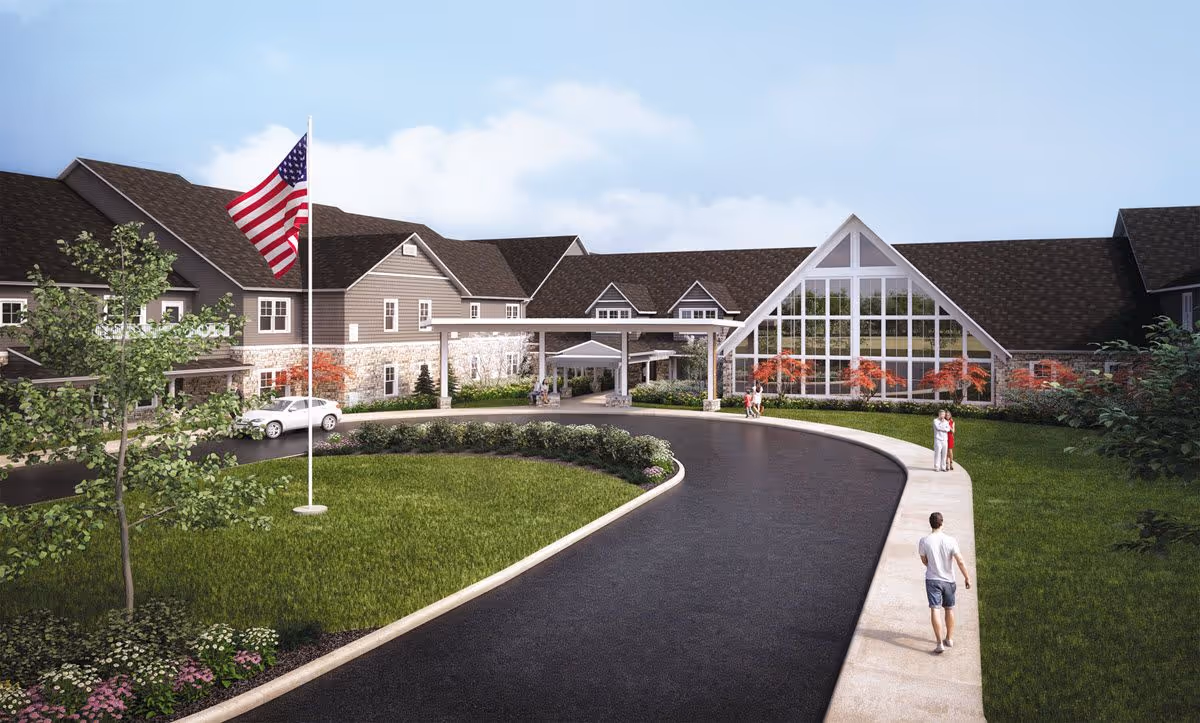 Front entrance of a senior living building with a circular driveway, American flag, a large glass atrium, and people walking on the sidewalk.
