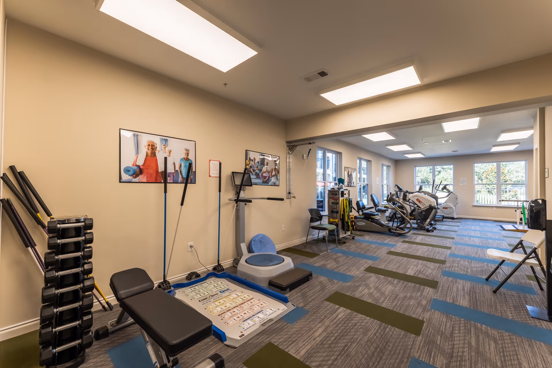 A well-lit fitness room with exercise equipment including a weight bench, dumbbells, resistance bands, stationary bikes, and other workout machines. The room has large windows letting in natural light and posters of elderly people exercising on the walls.