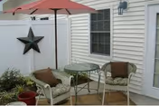 Small outdoor patio with two wicker chairs and cushions, a round glass-top table beneath a red umbrella, potted plants, and a decorative star on white siding.