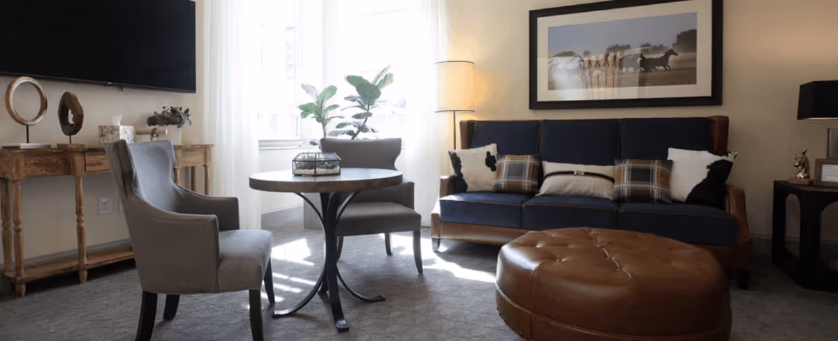 A cozy living room with a blue sofa adorned with patterned pillows, a round brown leather ottoman, two gray upholstered chairs around a small round table, a wooden console table with decorative items, a floor lamp, and a framed picture of horses on the wall. Natural light comes through a window with sheer white curtains.