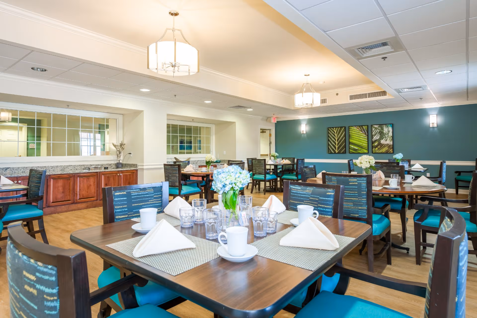 A bright and clean dining room with multiple wooden tables and chairs featuring teal cushions. Each table is set with white napkins, cups, glasses, and a centerpiece of flowers. The room has light-colored walls with a teal accent wall adorned with three framed leaf artworks. Ceiling lights and wall sconces provide illumination.