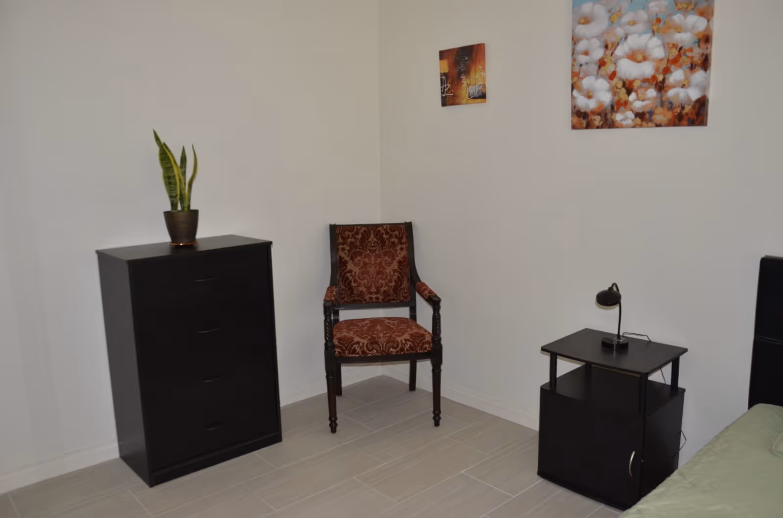 Corner of a simple bedroom with a dresser topped by a potted plant, an upholstered chair, a nightstand with a lamp, and floral wall art.