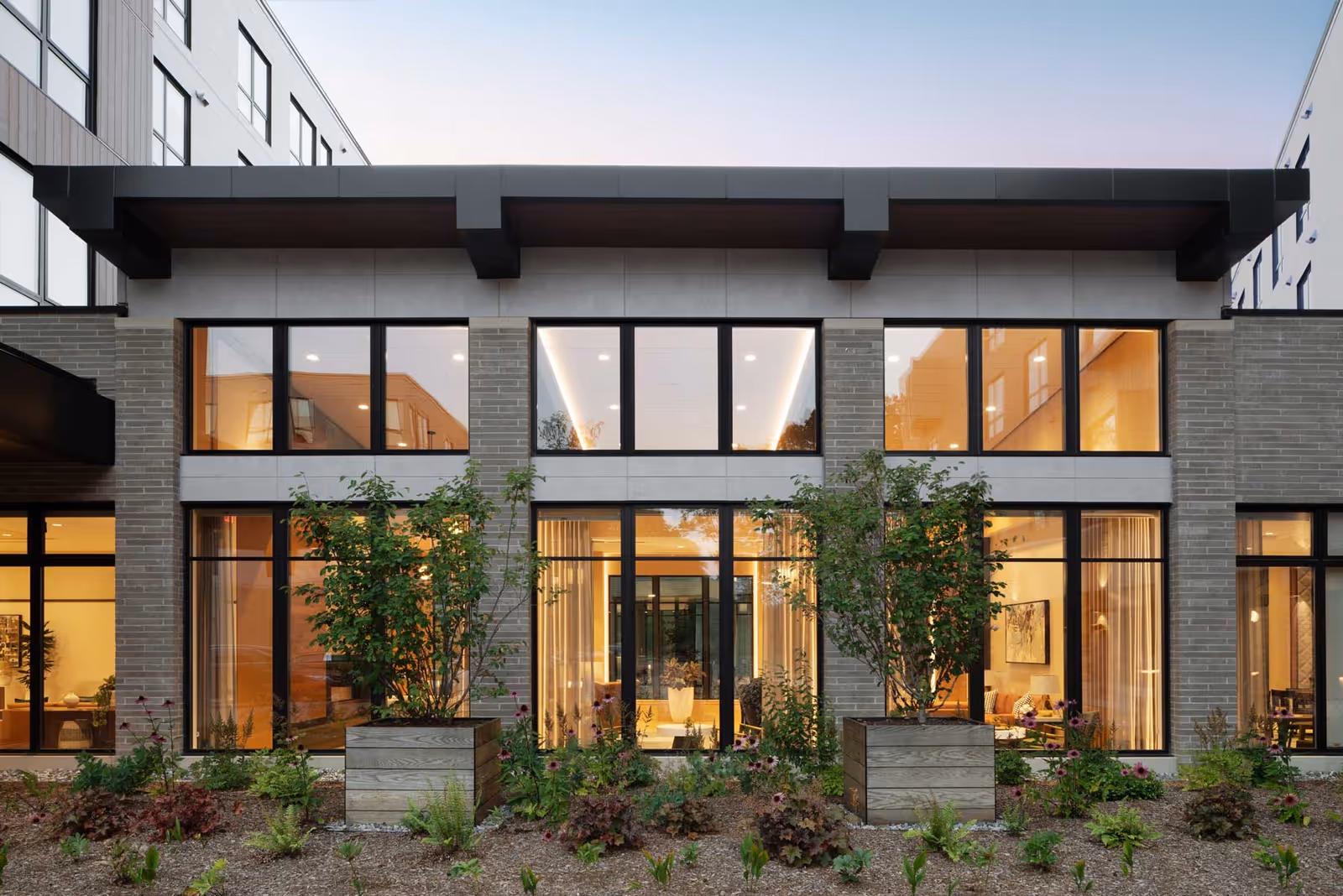 Exterior view of a modern senior living facility building with large windows showing warm interior lighting and greenery in front, including two large planter boxes with bushes and various plants in the garden bed.