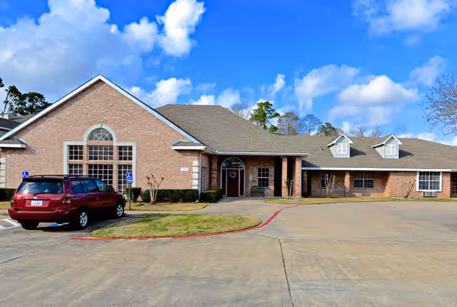 Exterior view of a single-story brick building with a pitched roof under a blue sky with some clouds. There is a red SUV parked in a handicapped parking space near the entrance, which has a covered porch area with columns. The building has large windows and a concrete driveway in front.