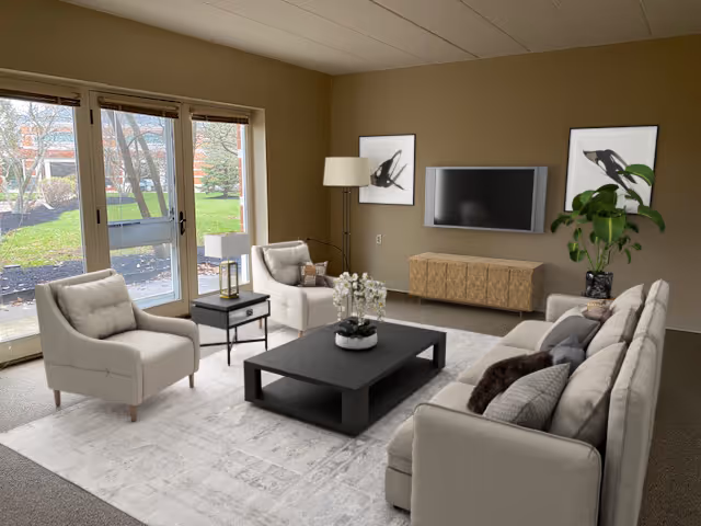 A cozy living room with beige walls and carpeted floor featuring a large beige sofa with multiple pillows, two matching armchairs, a black coffee table with a white flower arrangement, a side table with a lantern, a floor lamp, a wall-mounted flat-screen TV, two framed abstract artworks, and a large potted plant. Large glass doors open to a green outdoor area.