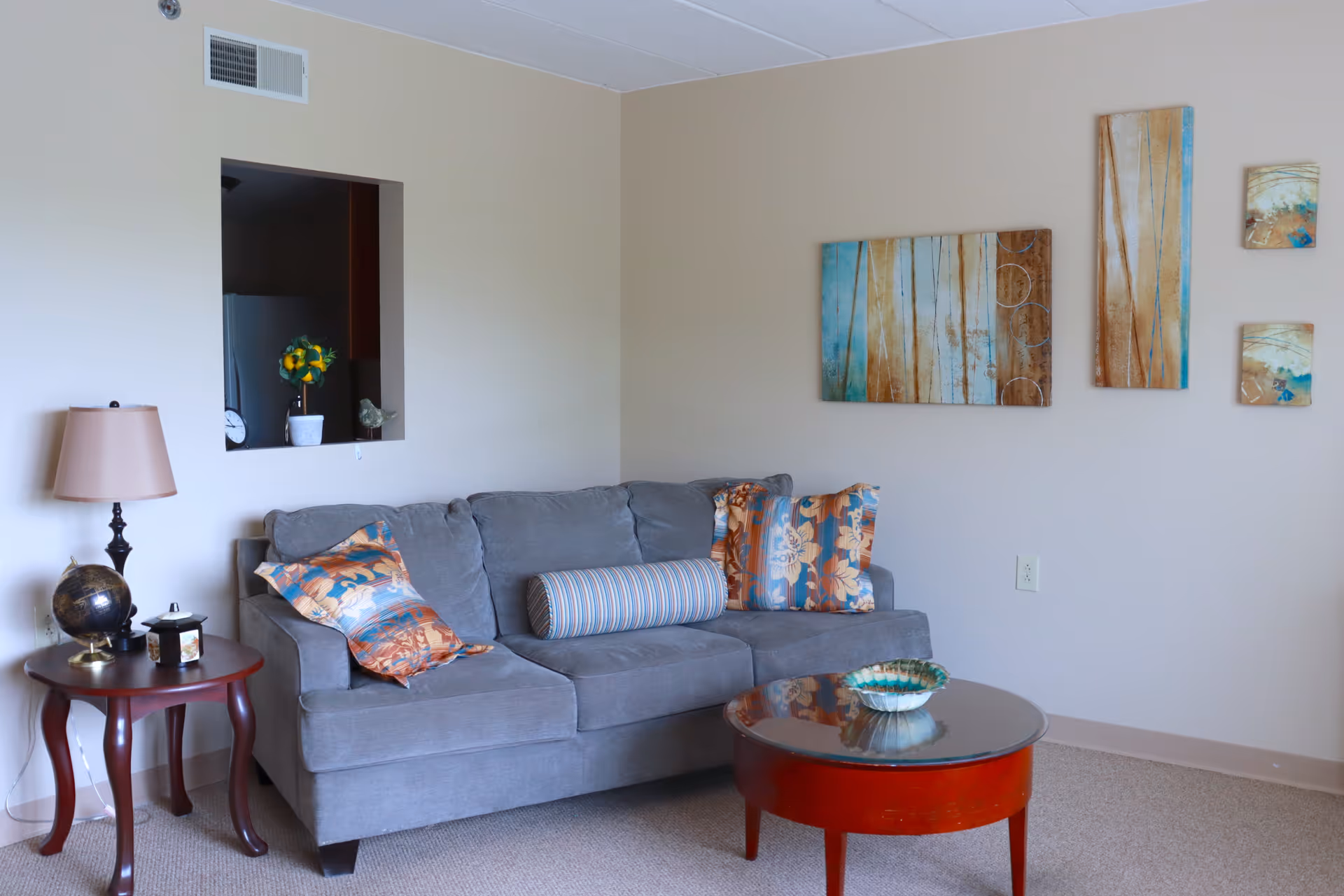 A cozy living room with a gray sofa adorned with colorful patterned pillows and a striped bolster pillow. A round wooden coffee table with a decorative bowl sits in front of the sofa. To the left, there is a small wooden side table with a lamp, a globe, and a small decorative item. The wall behind the sofa features a set of four abstract paintings in shades of blue, beige, and brown. A small open window in the wall reveals a glimpse of a kitchen area with a plant and a clock.