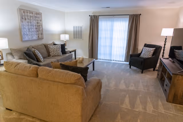Carpeted living room with two sofas, two armchairs, a coffee table, TV stand, lamps and a sliding glass door with curtains.