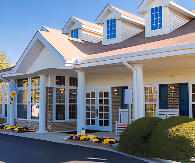 Front exterior view of a senior living facility building with white columns, large windows, a covered entrance, and a well-maintained garden with yellow flowers and green bushes under a clear blue sky.