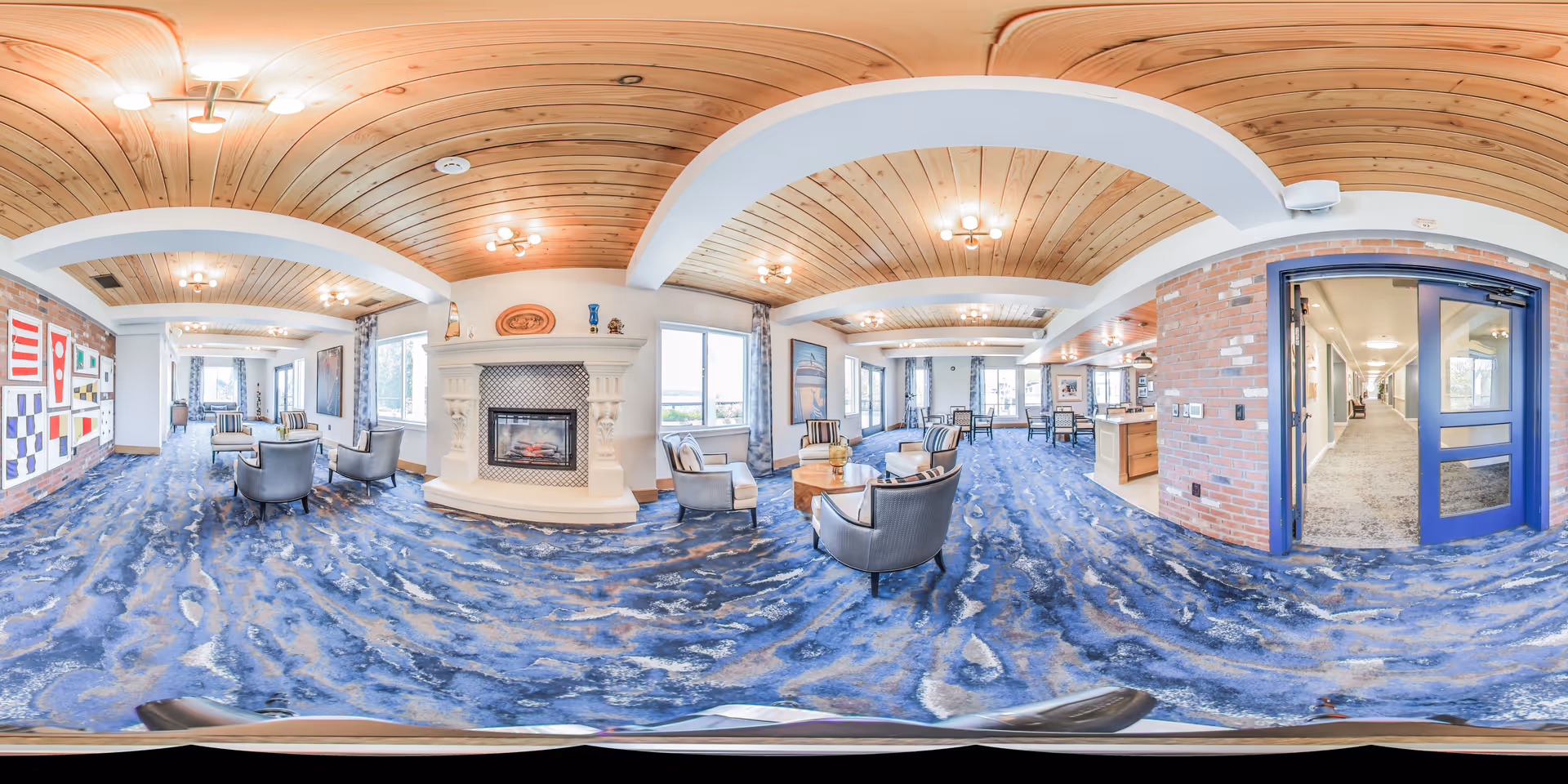 A spacious and well-lit senior living common area with blue patterned carpet, wooden ceiling with white beams, and multiple seating arrangements including armchairs and small tables. There is a white fireplace in the center, large windows letting in natural light, and a hallway visible through a glass door on the right side. The walls feature brick accents and nautical-themed decorations.