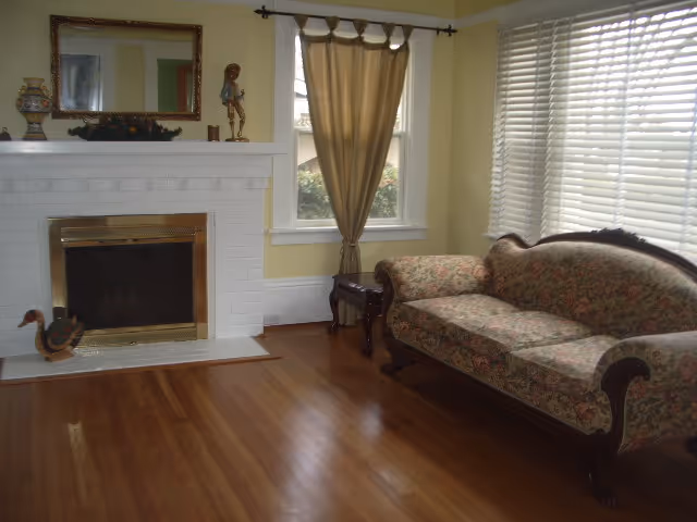 A cozy living room with a floral upholstered vintage sofa, a white brick fireplace with a brass frame, a large mirror above the fireplace, decorative items on the mantel, a window with beige curtains, and wooden flooring.