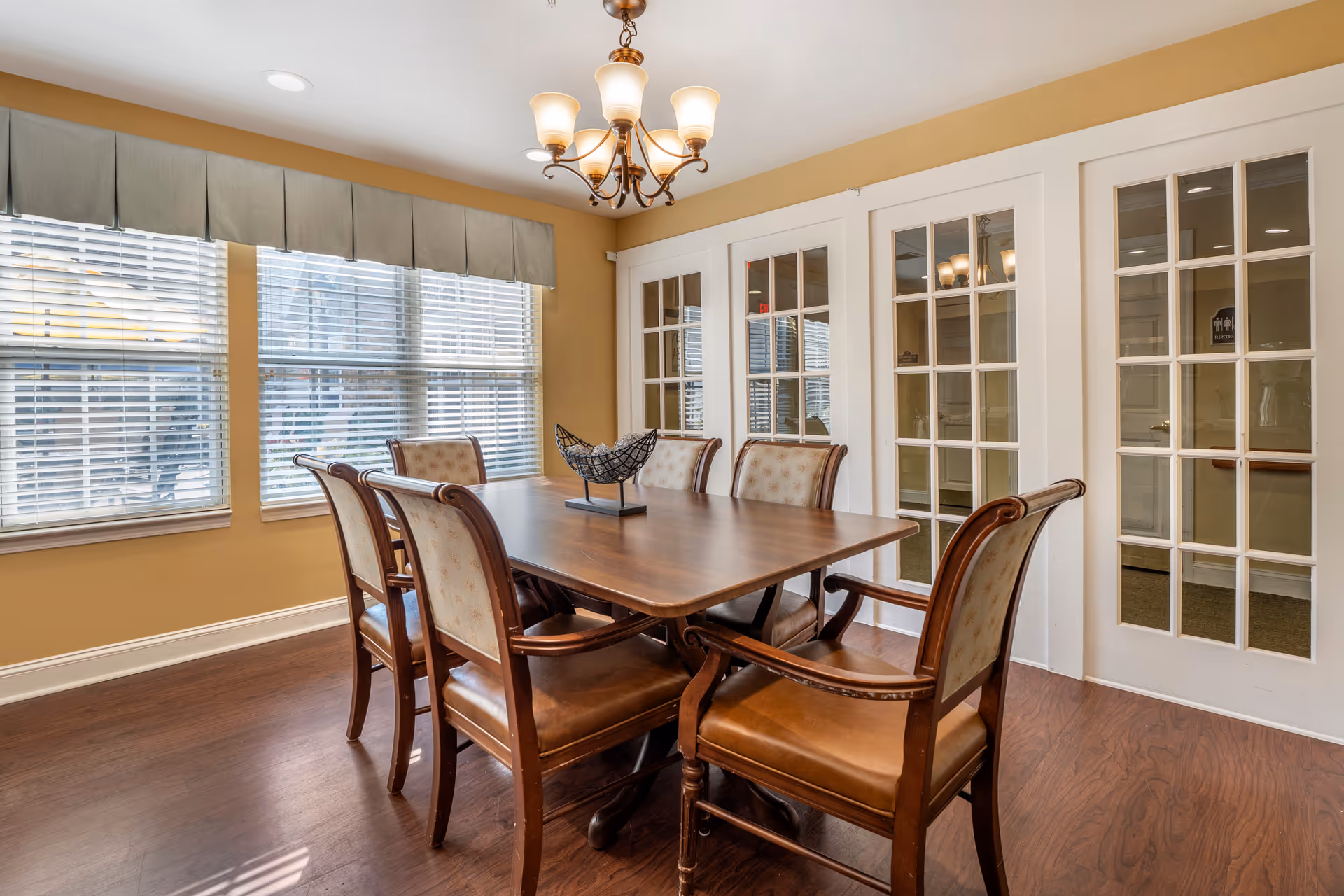 A dining room with a wooden rectangular table surrounded by six upholstered chairs. The room has large windows with blinds and a valance, a chandelier hanging from the ceiling, and white French doors with glass panes.