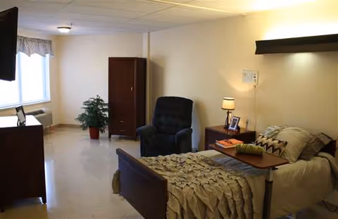A well-lit bedroom in a senior living facility featuring a hospital-style bed with beige bedding and a wooden headboard. Next to the bed is a wooden nightstand with a lamp, a framed photo, and some books. A dark upholstered recliner chair is positioned near a wooden wardrobe. A potted plant is placed near a window with a valance, and a wall-mounted TV is visible on the left side.