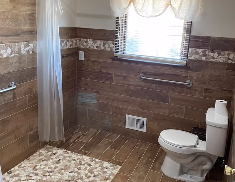 Accessible bathroom with tan tile walls, shower curtain, toilet, grab bars, and window.