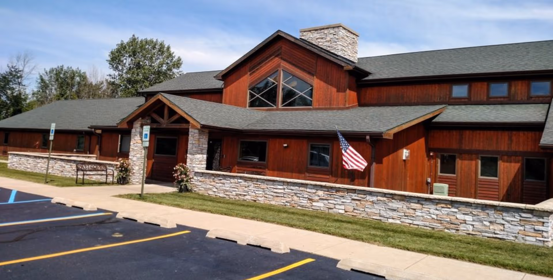 Exterior view of a single-story assisted living facility with wooden siding and stone accents. The building has a pitched roof with a chimney and large windows. There is a small stone wall in front of the building, an American flag mounted on the wall, and a parking lot with marked spaces including handicapped spots.