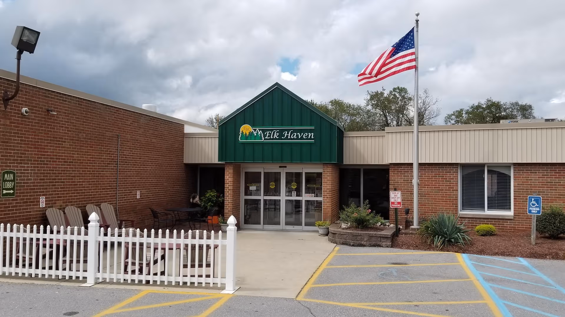 Front entrance of Elk Haven Nursing Home featuring a green awning with the facility's name, an American flag on a flagpole, a white picket fence enclosing a small seating area with chairs and tables, and a parking area with handicap parking spaces.