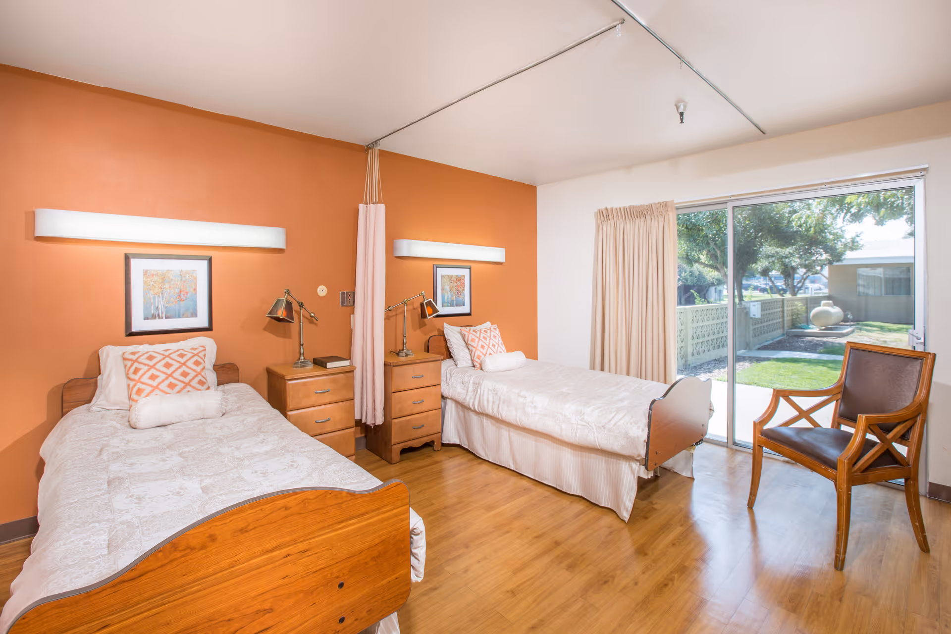 A bright and clean bedroom with two single beds, each with white bedding and decorative pillows. Between the beds are two wooden nightstands with lamps and a book. The room has an orange accent wall with framed artwork above each bed. A curtain divider hangs from the ceiling between the beds. There is a wooden chair with a cushioned seat near a large sliding glass door that opens to a green outdoor area with trees and a decorative pot.