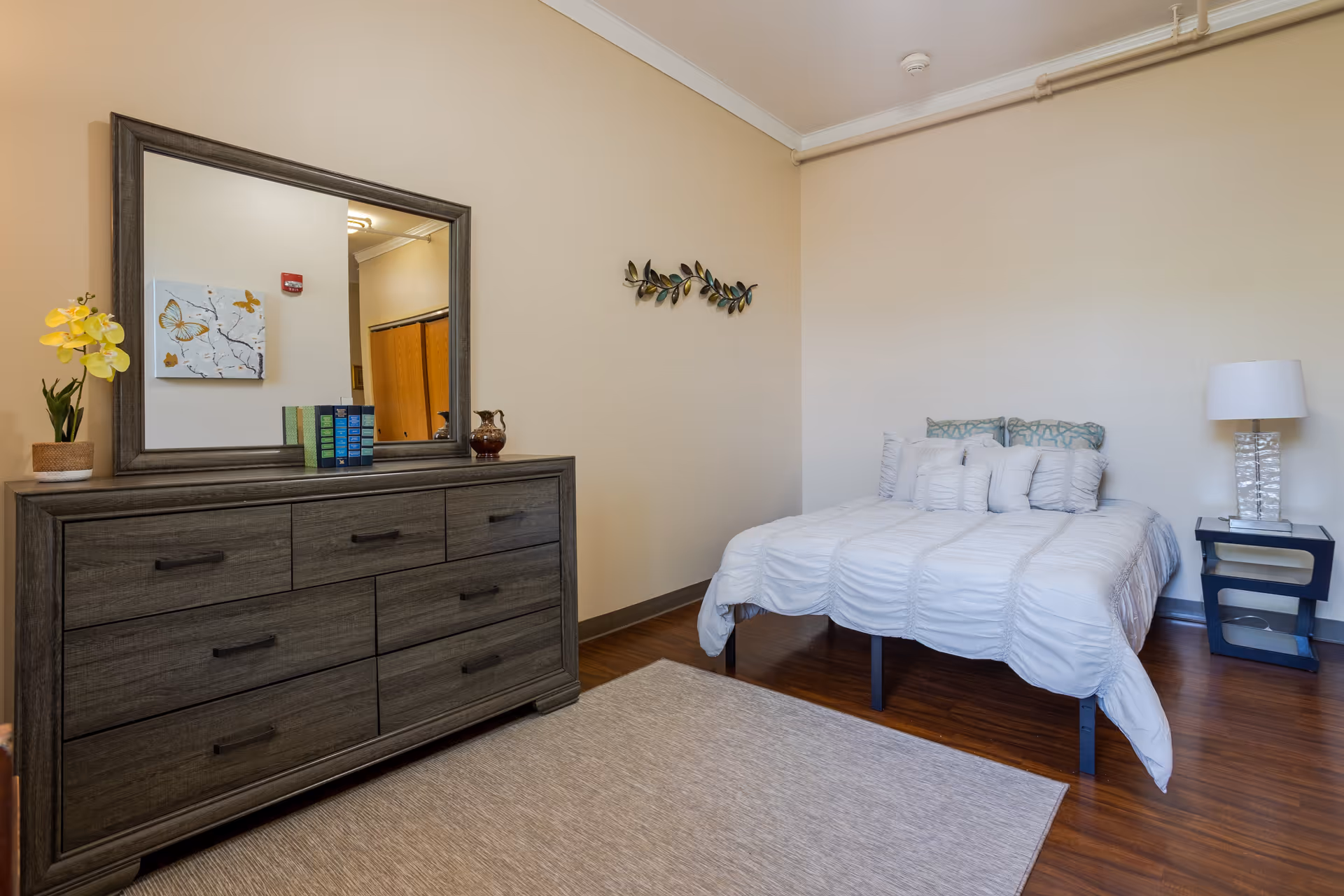 A neatly arranged bedroom with a bed covered in white bedding and multiple pillows. Next to the bed is a modern nightstand with a clear glass lamp. Across from the bed is a dark wooden dresser with six drawers, topped with a large mirror, a potted yellow orchid, a small decorative pitcher, and a few books. The room has light beige walls, wooden flooring, and a light-colored area rug.