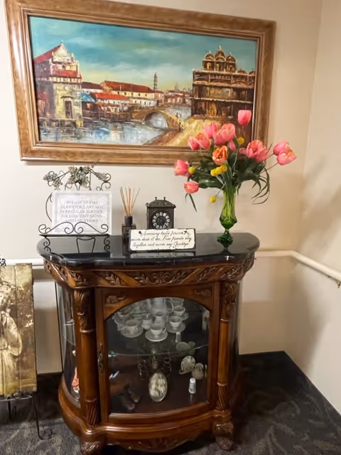 A decorative wooden cabinet with glass doors displaying glassware and decorative items inside. On top of the cabinet, there is a green vase with pink and yellow tulips, a small clock, reed diffuser sticks, and two small signs with text. Above the cabinet hangs a framed painting depicting a historic architectural scene with buildings and a bridge over water.