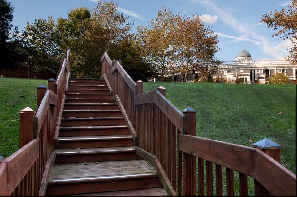 Wooden stairs with railings leading up a grassy hill towards a building with a dome and columns, surrounded by trees under a blue sky with some clouds.