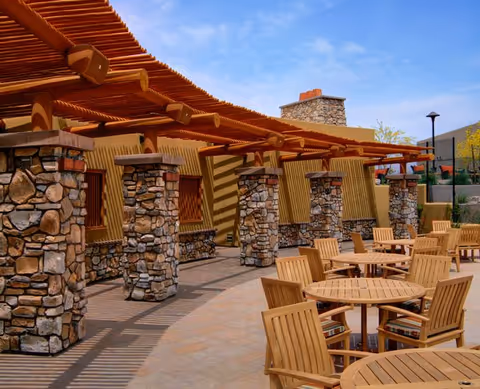 Outdoor patio with round wooden tables and chairs under a pergola supported by stone columns.