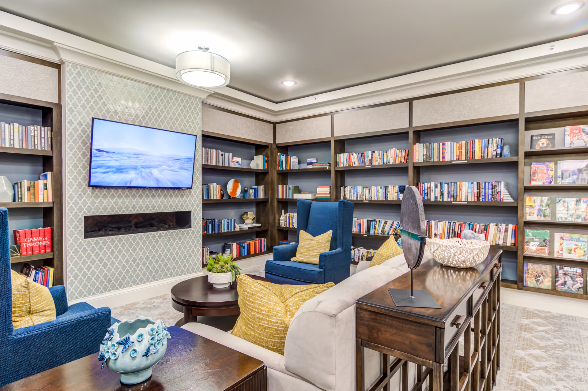 A cozy library room with built-in dark wood bookshelves filled with books and magazines. The room features a wall-mounted flat screen TV above a modern fireplace with a patterned tile surround. There are comfortable blue armchairs with yellow pillows and a beige sofa with yellow pillows arranged around a round wooden coffee table. Decorative items are placed on a wooden console table behind the sofa. The room has a soft, neutral color palette and recessed lighting in the ceiling.